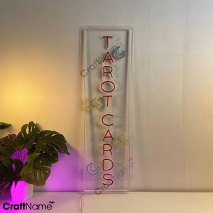 May include: A clear acrylic sign with the word "TAROT CARDS" spelled vertically in red neon lettering. The sign is decorated with small yellow stars and blue crescent moons.