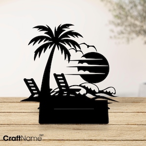 May include: A black metal silhouette sign featuring a tropical scene with a palm tree, two lounge chairs, a large sun, and stylized ocean waves. The design is mounted on a rectangular base, creating a decorative display piece for a flat surface.