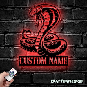 May include: A black metal wall sign featuring a coiled cobra with an open mouth and fangs, illuminated by a red LED glow. Below the snake is a rectangular plaque with the text CUSTOM NAME. A small remote control is held in the foreground.