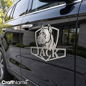 Op de afbeelding: Een metallic zilveren auto-embleem met een gestileerde hondenkop in een schildvorm en de naam JACK in dikke hoofdletters in het midden. Het embleem is bevestigd op de zijdeur van een glanzend zwart voertuig.