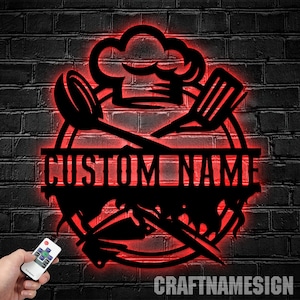 May include: Black metal silhouette of a chef's hat, crossed kitchen utensils, and a banner with the text "CUSTOM NAME". The silhouette is illuminated with red LED lights against a brick wall background.