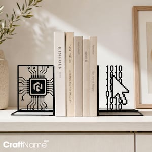 May include: A pair of black metal bookends supporting four books. One bookend features a central computer chip design with circuit board lines. The other bookend displays a pixelated computer mouse cursor icon surrounded by binary code digits 1 and 0.