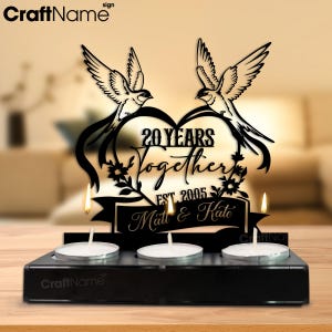Puede incluir: Portavelas de metal negro con tres velas de t&eacute;. La pieza central presenta un dise&ntilde;o en forma de coraz&oacute;n con dos p&aacute;jaros, el texto "20 YEARS Together EST. 2005 Matt & Kate". Un art&iacute;culo decorativo para un aniversario de bodas.