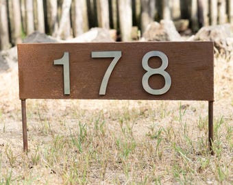 May include: A rectangular house number sign with a textured, rusted brown finish featuring the silver-colored metal numbers 178. The sign is mounted on two thin metal stakes that extend downward from the bottom corners of the rectangular plate.