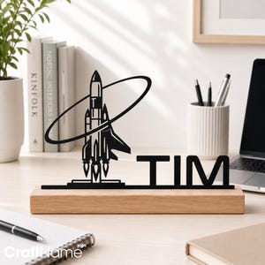 May include: A black metal desk sign featuring a rocket ship silhouette surrounded by an orbital ring, mounted on a rectangular light wood base. The name TIM is cut out in bold black capital letters next to the rocket ship design.