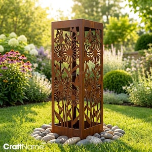 May include: A tall, rectangular, rust-colored metal garden sculpture featuring laser-cut designs of daisies and butterflies on its sides. The structure is positioned upright on a bed of smooth, light gray river stones set in green grass.