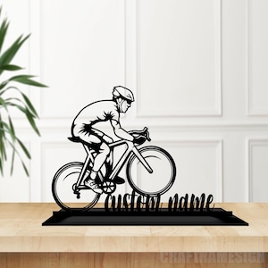 May include: A black silhouette cutout of a cyclist riding a road bike, mounted on a rectangular base. The design features a person wearing a helmet and cycling gear, positioned above the text custom name in a cursive script font.