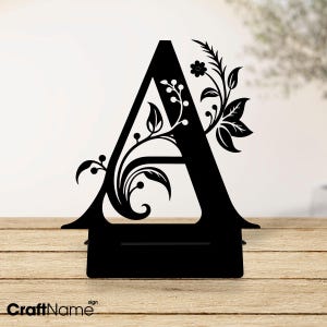 Puede incluir: Letra A de metal negro con un diseño floral. La letra está sobre una superficie de madera. Cartel CraftName.