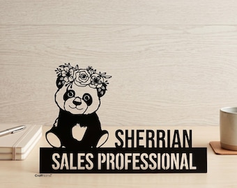 Op de afbeelding: Een zwart bureau-naambordje met een uitgesneden panda die een bloemenkrans draagt. Het bordje toont de tekst SHERRIAN en SALES PROFESSIONAL in dikgedrukte hoofdletters. Het ontwerp is gemaakt van stevig materiaal met een matte afwerking.