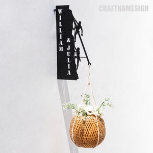 Puede incluir: Un letrero de pared de metal negro con los nombres "William & Julia" en letras blancas. El letrero representa dos siluetas de personas escalando. Una maceta colgante con flores blancas y una cesta de mimbre marrón cuelga del letrero.