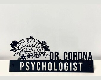 May include: A black metal desk sign featuring a cutout of a human brain surrounded by flowers. The text reads DR. CORONA on the top right and PSYCHOLOGIST in a rectangular base at the bottom. The design is flat and monochromatic.