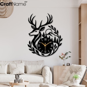 May include: A black wall clock featuring a laser-cut silhouette of a deer head, pine trees, and a mountain range. The circular clock face displays Roman numerals and gold-colored hands. The design is framed by stylized leaves and branches.
