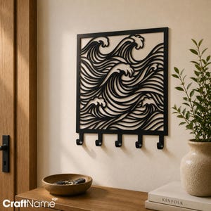 May include: A black metal wall-mounted organizer featuring a square frame with a cut-out design of stylized ocean waves. The piece includes five downward-facing hooks along the bottom edge for hanging items, constructed from solid metal.