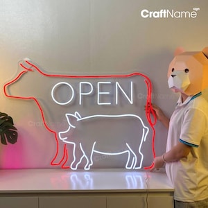 May include: White neon sign with the word "OPEN" in a large font. The sign is shaped like a cow with a pig inside the cow. The cow is outlined in red neon.