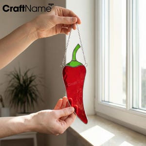 May include: A red stained glass chili pepper with a green stem, hanging from a silver chain. The pepper is held up by a person's hands. The text "CraftName" is in the upper left corner and "CraftName" is on the pepper.