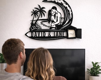 Può includere: Una decorazione da parete in metallo nero che raffigura la silhouette di una coppia che si bacia all'interno di una grande onda, con palme e una spiaggia. La base a forma di tavola da surf reca il testo DAVID & JANE e una piccola scatola.