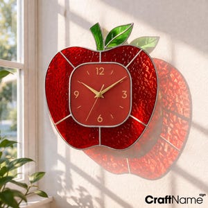 May include: A red apple-shaped wall clock with a square clock face. The clock has gold hands and a red background. The apple is made of textured red glass with a green glass leaf and stem. The clock is mounted on a beige wall.