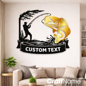 May include: A black metal wall sign featuring a silhouette of a person fishing under a tree, with a large gold metallic fish jumping out of the water. A black banner at the bottom contains the white text CUSTOM TEXT in a bold sans-serif font.