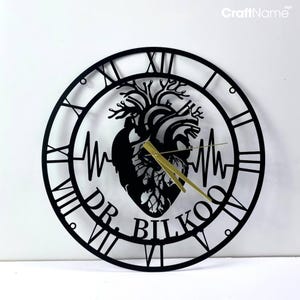 Op de afbeelding: Zwarte metalen wandklok met Romeinse cijfers. De wijzerplaat heeft een gestileerd anatomisch hart met een hartslaglijn en de tekst "DR. BILKO" eronder.