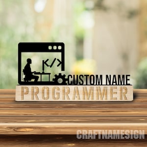 May include: A black acrylic cutout featuring a silhouette of a person sitting at a computer desk with code brackets and a gear icon. The text CUSTOM NAME is displayed next to the silhouette, and the word PROGRAMMER is engraved on a wooden base.