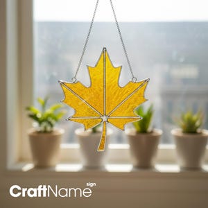 May include: A yellow stained glass maple leaf suncatcher with a textured surface and silver metal soldering. The leaf is suspended by a thin silver chain, featuring a distinct central vein and pointed lobes, designed to hang in a window.