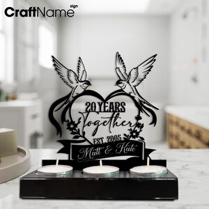 Puede incluir: Portavelas de madera negra con tres velas de t&eacute;. La pieza central presenta un dise&ntilde;o en forma de coraz&oacute;n con dos p&aacute;jaros, el texto "20 YEARS Together EST. 2005" y los nombres "Matt & Kate".