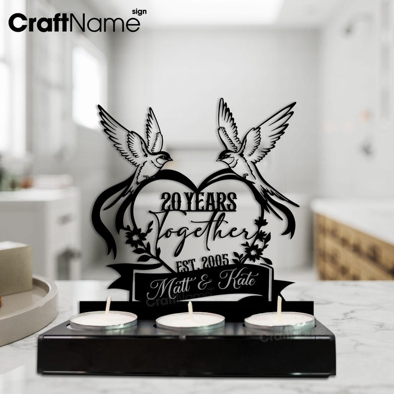 Puede incluir: Portavelas de madera negra con tres velas de t&eacute;. La pieza central presenta un dise&ntilde;o en forma de coraz&oacute;n con dos p&aacute;jaros, el texto "20 YEARS Together EST. 2005" y los nombres "Matt & Kate".