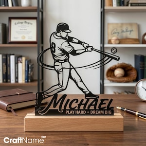 Peut inclure: Une silhouette en métal noir représentant un joueur de baseball en train de frapper, montée sur un socle en bois rectangulaire. Le design affiche le nom MICHAEL et le texte PLAY HARD • DREAM BIG. Le joueur porte un maillot avec le numéro 8.