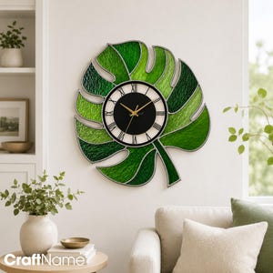 May include: A wall clock shaped like a large monstera leaf with textured green glass panels and a central circular face. The clock face features a black background, white border, black Roman numerals, and thin gold-coloured hour and minute hands.