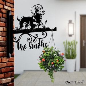 May include: Black metal sign with a dachshund silhouette and the text "The Smiths" hanging on a brick wall. A hanging planter with colorful flowers is attached to the sign.
