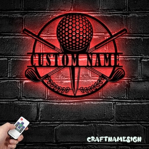 May include: A circular black metal wall sign featuring a golf ball on a tee, crossed golf clubs, and a laurel wreath design. The sign includes a horizontal bar with the text CUSTOM NAME and is illuminated by a red LED light glow.