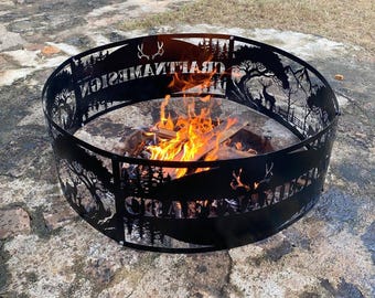 May include: A circular black metal fire pit ring featuring laser-cut silhouettes of deer, pine trees, and mountains. The text CRAFTNAMESIGN is cut into the metal panels. A bright orange fire burns inside the ring on a stone surface.