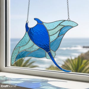 May include: A stained glass suncatcher shaped like a manta ray, featuring a deep blue central body and translucent light blue wings with textured glass panels. The piece is suspended by a thin metal chain, showcasing intricate leaded lines.