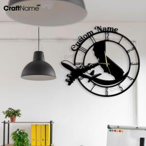 May include: Black metal wall clock with a plane flying over a state outline. The clock face has Roman numerals and the words "Custom Name" are printed above the clock face.
