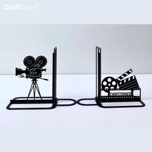 Peut inclure: Une paire de serre-livres en métal noir avec des silhouettes sur le thème du cinéma. L'un présente une caméra de film vintage sur trépied, l'autre une bobine, un clap de cinéma et une pellicule avec le nom CHRISTOPHER découpé au centre.