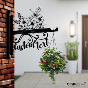 Può includere: Vaso da fiori appeso in metallo nero con un design di libellula e fiori. Il vaso ha uno spazio per il testo personalizzato e un gancio per appenderlo. Il vaso è mostrato con un cestino appeso di fiori.