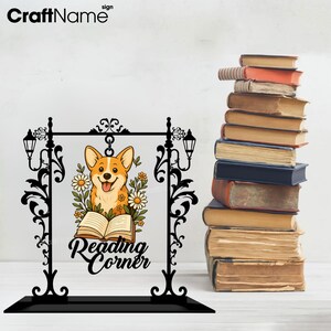 May include: A black metal sign with a corgi illustration, flowers, and the words "Reading Corner." The sign has a decorative frame with lamps and sits on a black base. A stack of old books is to the right.