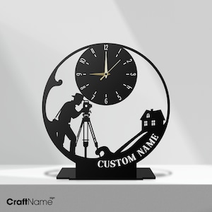 May include: A black circular desk clock featuring a silhouette of a surveyor using a theodolite on a tripod, a house, and the text CUSTOM NAME. The clock face has white numbers and thin gold hands, all mounted on a flat rectangular base.