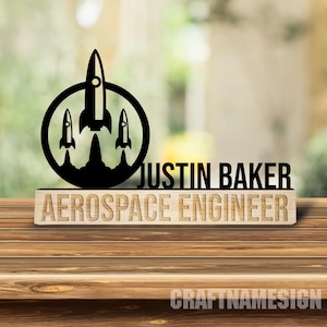 Op de afbeelding: Een zwart, lasergesneden bureau-ornament met een cirkelvormig embleem met drie opstijgende raketten, naast de tekst JUSTIN BAKER. Daaronder staat een lichtgekleurde houten voet met de gegraveerde tekst AEROSPACE ENGINEER.