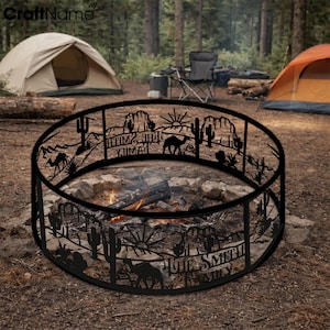 May include: A black metal fire pit ring with a desert landscape design, including cacti, camels, and the text "THE SMITH FAMILY". The fire pit is set up outdoors with a campfire burning inside, surrounded by rocks and trees.