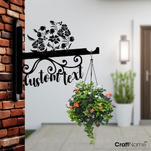May include: Black metal sign with a floral design and the text "custom text". A hanging planter with green, orange, and white flowers is attached to the sign.