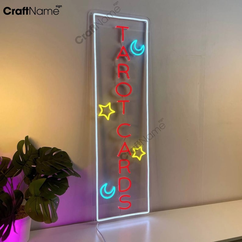 May include: A neon sign with the words "TAROT CARDS" in red letters. The sign is surrounded by a white frame and has yellow stars and blue crescent moons around the letters.