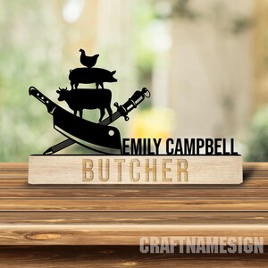 May include: A black metal cutout featuring a stacked chicken, pig, and cow silhouette above crossed butcher knives. The text EMILY CAMPBELL is displayed in black, while the word BUTCHER is engraved on a light-coloured wooden base block.