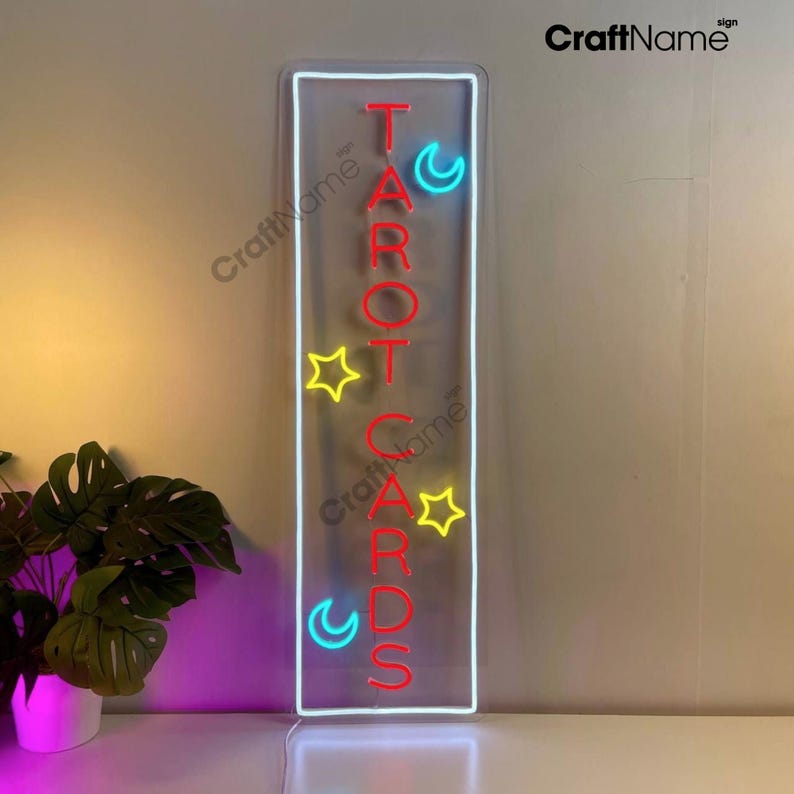 May include: Neon sign with the word "TAROT CARDS" in red letters, with a yellow star and blue crescent moon on either side of the letters. The sign is framed in white and has a clear background.