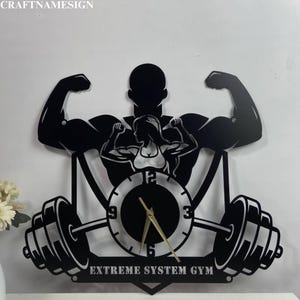 May include: Black metal wall clock shaped like a weightlifter lifting a barbell. The clock face is in the center of the design and features a gold-colored hour and minute hand. The clock face has black numbers and a black background. The text "EXTREME SYSTEM GYM" is printed below the clock face.