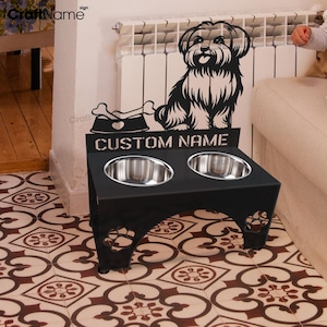 May include: Black metal dog bowl stand with two stainless steel bowls. The stand features a silhouette of a dog with a bone and the text "CUSTOM NAME".