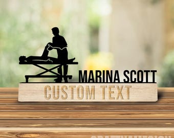 Könnte beinhalten: Eine schwarze Acrylsilhouette eines Massagetherapeuten, der an einem Kunden auf einer Liege arbeitet, montiert auf einem hellen Holzsockel. Der Text MARINA SCOTT ist schwarz und CUSTOM TEXT ist in den Holzsockel eingraviert.