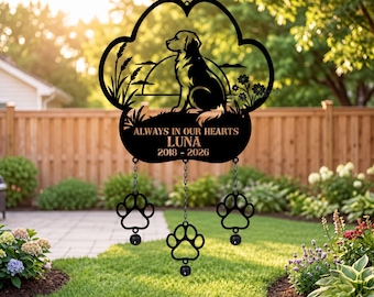 Könnte beinhalten: Ein schwarzes Metall-Windspiel in Form eines großen Pfotenabdrucks mit der Silhouette eines Hundes, hohem Gras und Blumen. Der Text lautet ALWAYS IN OUR HEARTS LUNA 2018 - 2026. Drei kleinere Pfotenanhänger hängen an Ketten darunter.
