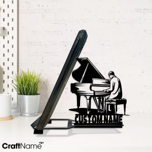 May include: Black metal phone stand featuring a piano player silhouette and a grand piano. The stand has a slot to hold a phone and includes the text "CUSTOM NAME". The brand name "CraftName" is also visible.