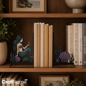 May include: A pair of black metal bookends featuring a mermaid with long teal hair, a purple seashell top, and a teal tail holding a small purple starfish, alongside a second bookend with a large purple seashell and teal seaweed accents.